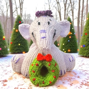 May include: A knitted white elephant toy with purple accents, wearing a purple hat and a green wreath with a red bow. The elephant is sitting in front of Christmas trees.