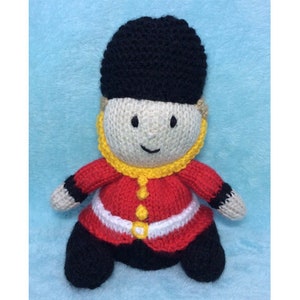 May include: A knitted toy soldier wearing a red and white uniform with a black hat. The toy has a yellow collar and a friendly face.