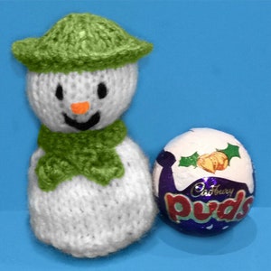 May include: A handmade knitted snowman with a green hat and scarf, next to a Cadbury Puds chocolate egg. The snowman has a white body, black eyes, and an orange nose. The chocolate egg is wrapped in blue and white foil.