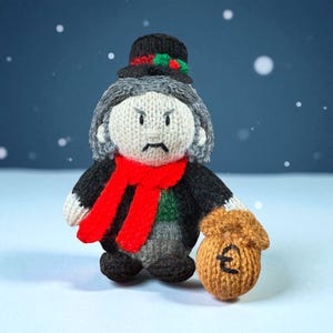 May include: Hand-knitted Ebenezer Scrooge doll with a stern expression, gray hair, and a red scarf. He wears a black top hat with holly and a black coat over a green vest. He clutches a brown money bag with a pound symbol.