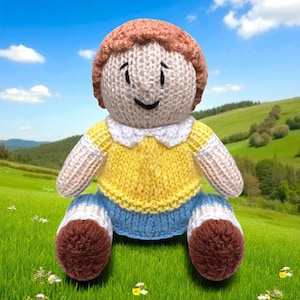 May include: A handmade knitted doll with a smiling face, wearing a yellow shirt with a white collar, blue pants, and brown shoes. The doll has brown hair and is sitting on a green grassy hill with a blue sky background.