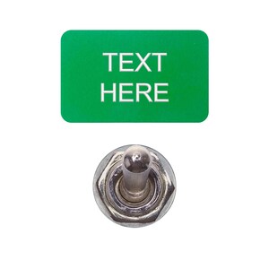 以下が含まれることがあります： 白い文字で「TEXT HERE」と書かれた緑色の長方形のボタン。ボタンは銀色のトグルスイッチに取り付けられています。
