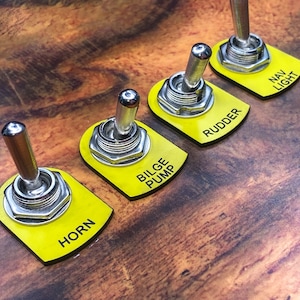 Pode incluir: Cinco interruptores de alavanca amarelos com letras pretas. Os interruptores são etiquetados como "Horn", "Bilge Pump", "Rudder", "Nav Light".