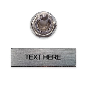 May include: A silver toggle switch with a round knob and a rectangular metal plate with the text "TEXT HERE" engraved on it.