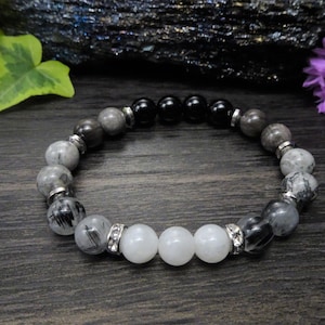 May include: A black, grey, and white beaded bracelet with silver accents. The bracelet features a variety of round beads, including black onyx, clear quartz, and grey agate.