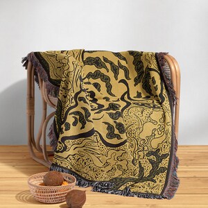 Pu&ograve; includere: Una coperta in tessuto intrecciato con un motivo astratto nero e verde su uno sfondo giallo senape. La coperta &egrave; drappeggiata su una sedia in vimini e ha frange sui bordi.