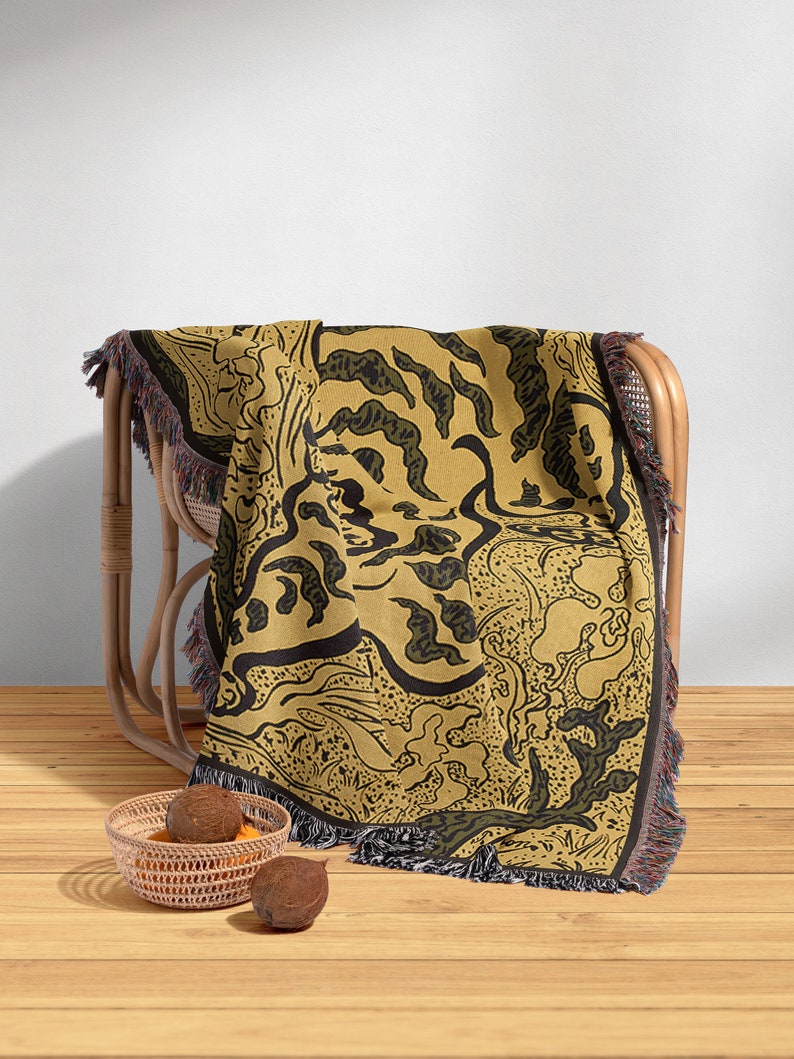 Pu&ograve; includere: Una coperta in tessuto intrecciato con un motivo astratto nero e verde su uno sfondo giallo senape. La coperta &egrave; drappeggiata su una sedia in vimini e ha frange sui bordi.