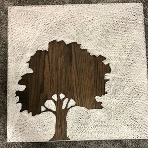 May include: A square wooden art piece featuring a tree silhouette. The tree is cut out of dark brown wood, and the background is filled with white string art, creating a textured effect. The artwork is displayed on a gray surface.