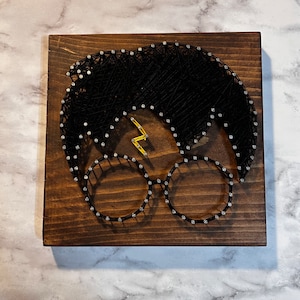 May include: String art portrait of a character with black hair, round glasses, and a lightning bolt scar. The design is created with black thread on a stained wooden board. The glasses and scar are accented with silver and gold colored nails.