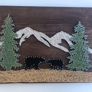 May include: A wooden string art piece depicting a mountain scene with two bears. The mountains are white, the trees are green, and the bears are black. The background is a dark brown, and the ground is tan.