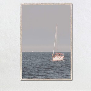 Puede incluir: Un velero blanco con una franja roja navega en un mar azul tranquilo bajo un cielo gris.