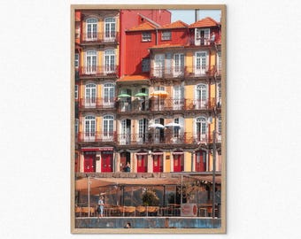 May include: A colorful building with red, yellow, and white accents. The building has multiple balconies with white railings and several windows. There are umbrellas on the balconies and a cafe in front of the building.