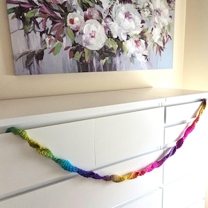 May include: A colorful crocheted garland with a rainbow of colors, including pink, purple, blue, green, yellow, and orange, is draped over a white dresser.