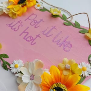May include: Pink wooden sign with the text "Pot it like it's hot" written in purple. The sign is decorated with artificial flowers, including sunflowers, daisies, and a bee.
