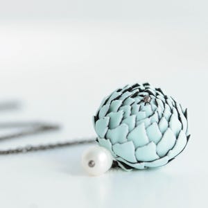 Può includere: Una collana con un ciondolo a forma di succulenta o carciofo azzurro con bordi scuri, accentuato da una singola perla bianca. Il ciondolo è fissato a una sottile catena di metallo scuro che si estende al di fuori dell'inquadratura.