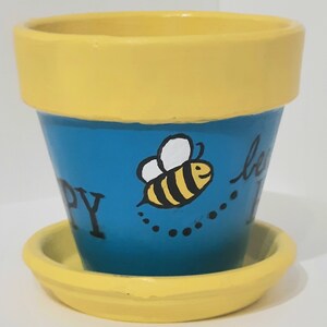 Puede incluir: Una maceta de cerámica amarilla y azul con un diseño de abeja y el texto "be happy". La maceta tiene un platillo amarillo a juego.