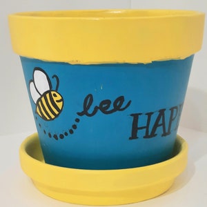 Puede incluir: Una maceta de cerámica amarilla y azul con un diseño de abeja y el texto "bee happy".