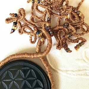 Könnte beinhalten: Eine braune Makramee-Kette mit einem schwarzen Steinanhänger, der ein geschnitztes Lebensblume-Design zeigt. Die Kette ist mit kleinen braunen und goldenen Perlen verziert.