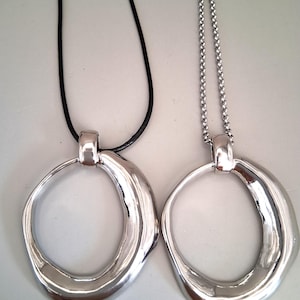 May include: Two silver-toned pendants with irregular, organic circular shapes. One pendant hangs from a smooth black cord, while the other is suspended from a silver-toned metal ball chain. Both pieces feature a polished, reflective finish.