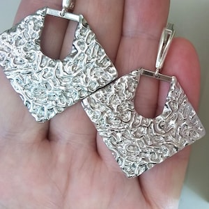 May include: A pair of silver earrings with a textured, geometric design. The earrings are square-shaped with a cutout in the centre.