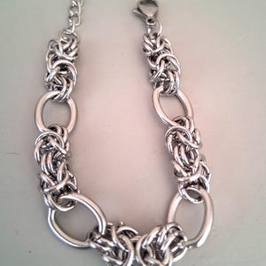 May include: A silver chain bracelet featuring a distinctive, intricate design. The bracelet is crafted from multiple linked oval shapes, each with a woven knot pattern.