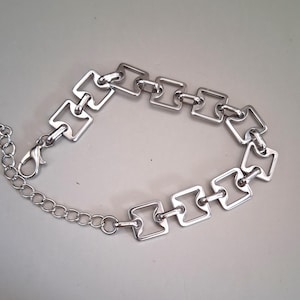 May include: A silver-toned metal bracelet featuring a series of interconnected square-shaped links with rounded inner corners. The bracelet includes a lobster claw clasp and an adjustable chain extension with circular links for a secure fit.