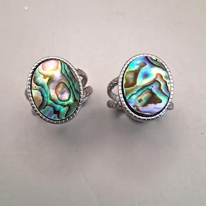 May include: Two silver rings featuring oval abalone shells. The shells show iridescent colours of green, blue, and gold. The rings have a textured band design and a silver bezel setting.