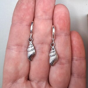 May include: A pair of silver seashell earrings. Each earring has a detailed, spiralled seashell pendant hanging from a hinged hoop. The earrings are made of a shiny, reflective metal, probably sterling silver, and are designed to be worn on the ear.