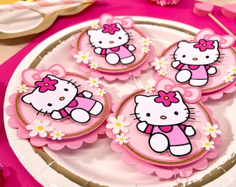 May include: Four pink and gold Hello Kitty cake toppers with white flowers. The toppers are on a white paper plate with a gold rim.
