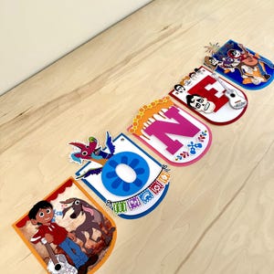 May include: A decorative banner spelling the word ONE, featuring layered cardstock cutouts with colorful illustrations of characters, a guitar, and festive patterns inspired by the movie Coco, including a blue letter O, pink N, and red E.