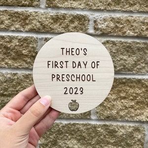 May include: A round wooden sign with the text "Theo's First Day of Preschool 2023" and a small apple graphic.