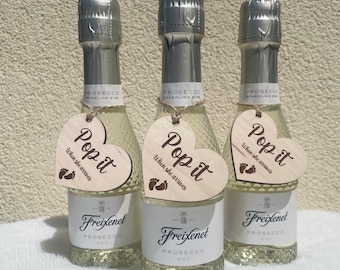 Puede incluir: Tres mini botellas de prosecco con etiquetas de madera en forma de corazón que dicen "Pop it when she arrives" con un símbolo de huella. El texto "Bottles Not Included" está en la parte inferior de la imagen.