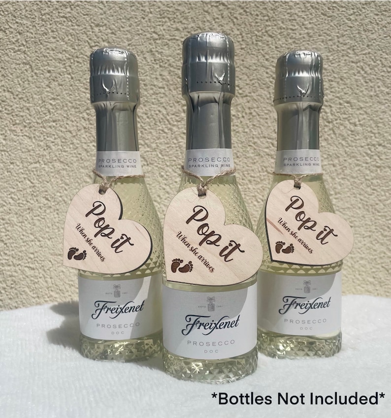 Op de afbeelding: Drie mini flessen prosecco met hartvormige houten labels waarop "Pop it when she arrives" staat met een voetafdruk symbool. De tekst "Bottles Not Included" staat onderaan de afbeelding.