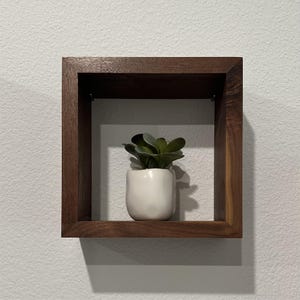 May include: A square wooden shelf with a small potted plant on it. The shelf is made of dark wood and has a simple design.