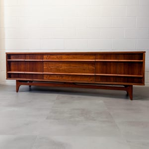 May include: A long, low-profile wooden sideboard featuring a rich brown walnut grain. The piece includes three central drawers stacked vertically and two side cabinets with horizontal shelving, all supported by four tapered wooden legs.