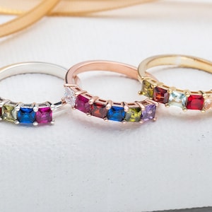 May include: Three rings with colorful gemstones. The rings are made of silver, rose gold, and gold. Each ring has seven square-shaped gemstones in a rainbow of colors.