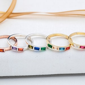 May include: Five rings with colorful rectangular gemstones. The rings are in rose gold, silver, and gold tones. Each ring features a different arrangement of gemstones in various colors, including red, purple, green, blue, and clear.