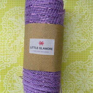 Pu&ograve; includere: Un rotolo di nastro di rete viola con un luccichio argentato. Il nastro &egrave; avvolto attorno a un tubo di carta marrone con un'etichetta che recita "LITTLE GLAMORE FOR EVERY OCCASION".
