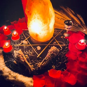 Pu&ograve; includere: Una scena mistica con una lampada di sale luminosa, candele rosse e petali di rosa sparsi. Due mazzi di salvia sono posti su una superficie scura, insieme a un calice d'argento e un disegno a forma di stella. L'atmosfera &egrave; arricchita da una luce soffusa.
