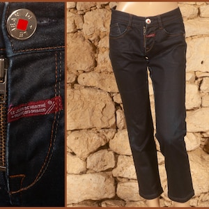 Puede incluir: Vaqueros de mezclilla azul oscuro con cremallera y cierre de botón. Los vaqueros tienen costuras naranjas y una etiqueta roja que dice "LE JEAN DE MARITHÉ + FRANÇOIS GIRBAUD". Los vaqueros se muestran en un maniquí.