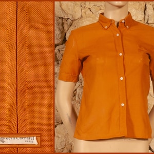 Peut inclure: Chemise orange à manches courtes avec boutons. La chemise a un col classique et une poche poitrine unique. Le tissu semble être un tissage texturé. L'étiquette indique "Jean Cacarel PARIS".