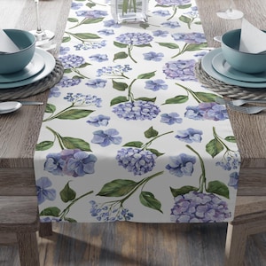 Könnte beinhalten: Ein weißer Tischläufer mit einem Aquarell-Blumenmuster aus blauen und lila Hortensien und grünen Blättern. Der Läufer liegt auf einem Holztisch mit Gedecken, darunter blaue Schalen, Teller und Weingläser.