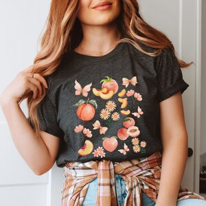 May include: A dark gray t-shirt with a peach design. The design features peaches, butterflies, flowers, and bows. The peaches are pink and orange with green leaves. The butterflies are blue and purple. The flowers are yellow and pink. The bows are pink.