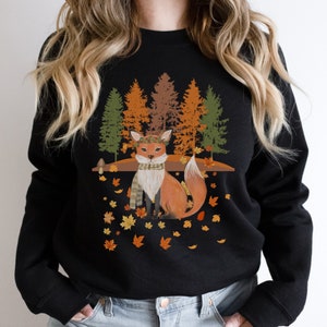 Puede incluir: Sudadera negra con una ilustraci&oacute;n de un zorro con tem&aacute;tica oto&ntilde;al. El zorro lleva una bufanda y una corona, rodeado de hojas ca&iacute;das y &aacute;rboles en tonos verdes, naranjas y marrones. El dise&ntilde;o evoca una est&eacute;tica oto&ntilde;al.