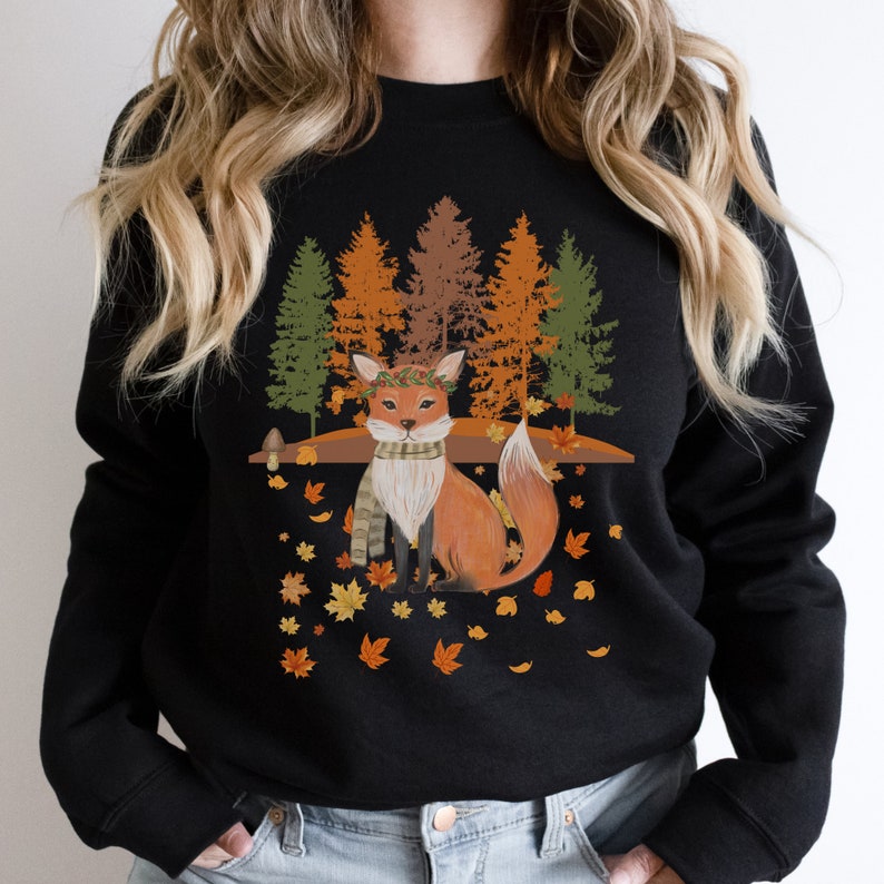 Puede incluir: Sudadera negra con una ilustraci&oacute;n de un zorro con tem&aacute;tica oto&ntilde;al. El zorro lleva una bufanda y una corona, rodeado de hojas ca&iacute;das y &aacute;rboles en tonos verdes, naranjas y marrones. El dise&ntilde;o evoca una est&eacute;tica oto&ntilde;al.