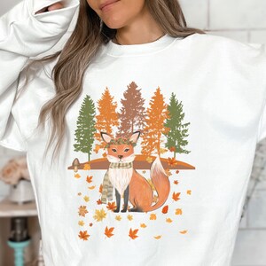 Puede incluir: Sudadera blanca con una ilustraci&oacute;n de un zorro en un entorno oto&ntilde;al. El zorro lleva una bufanda y una corona de hojas, con un fondo de &aacute;rboles y hojas cayendo. El dise&ntilde;o utiliza tonos naranja, marr&oacute;n y verde.
