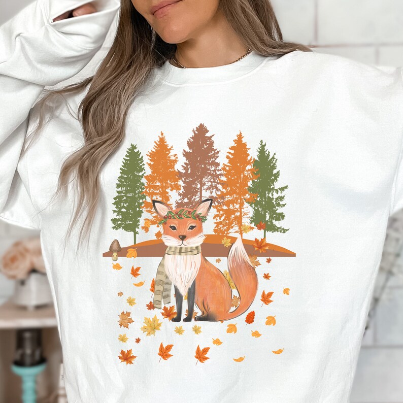 Puede incluir: Sudadera blanca con una ilustraci&oacute;n de un zorro en un entorno oto&ntilde;al. El zorro lleva una bufanda y una corona de hojas, con un fondo de &aacute;rboles y hojas cayendo. El dise&ntilde;o utiliza tonos naranja, marr&oacute;n y verde.