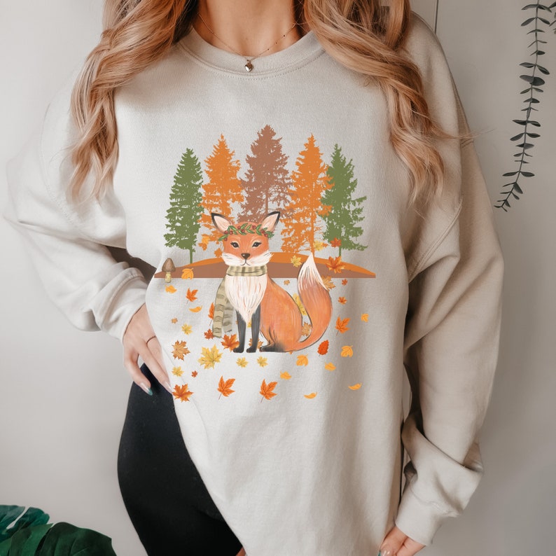 Puede incluir: Sudadera beige con una ilustraci&oacute;n de un zorro en una escena de bosque oto&ntilde;al. El zorro es naranja con una corona y bufanda verdes, rodeado de hojas ca&iacute;das y &aacute;rboles coloridos. Un dise&ntilde;o acogedor con tem&aacute;tica oto&ntilde;al.