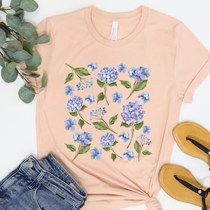 May include: A peach-coloured t-shirt with a floral print featuring blue hydrangeas and green leaves.