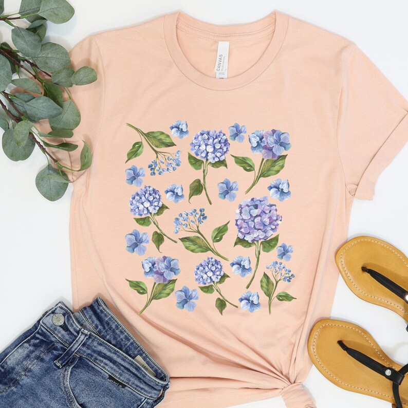 May include: A peach-coloured t-shirt with a floral print featuring blue hydrangeas and green leaves.